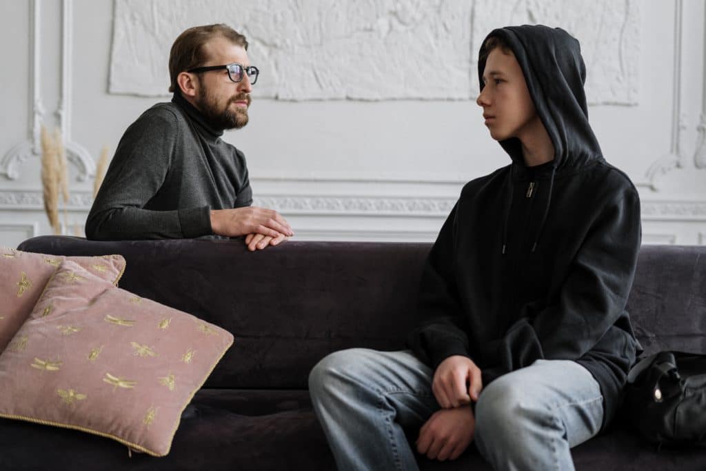 man in black jacket sitting on couch talking to teenage boy wearing a black hoodie
