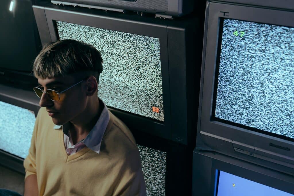 Man wearing glasses and brown button up shirt in front of multiple screens showing static