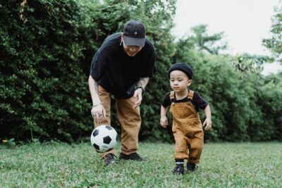 Man in black shirt helping a young boy in overalls play soccer